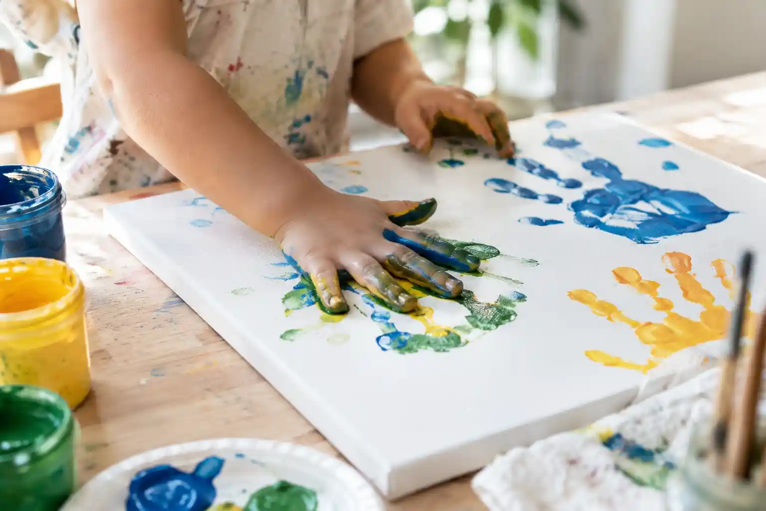 Small hands mid-handprint-painting on a canvas at a craft table